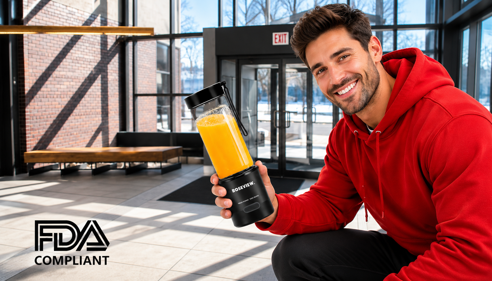 A smiling man in a red hoodie kneels in a modern glass-walled lobby while holding a black NutriBullet blender bottle filled with orange juice. The FDA Compliant logo is visible in the bottom-left corner.